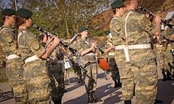Eine Gruppe von Soldaten in Tarnuniformen, die draußen Instrumente spielen, einige halten Klarinetten und andere Trommeln, stehen in Formation unter einem klaren Himmel.