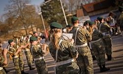 Eine Militärkapelle junger Menschen in grünen Uniformen spielt auf der Straße. Sie halten verschiedene Musikinstrumente in den Händen.