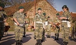 Eine Gruppe von Soldaten in Tarnuniformen steht im Freien in einer Reihe. Einige halten Musikinstrumente, und eine Person hält ein Dokument. Sie scheinen sich auf eine Zeremonie vorzubereiten.