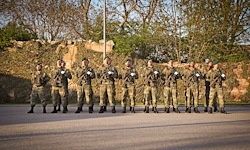 Eine Gruppe Soldaten in Tarnuniformen steht in einer Reihe auf einer Straße und hält Waffen. Bäume und eine Steinmauer sind hinter ihnen.
