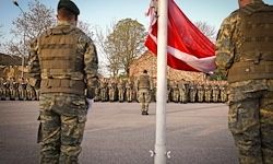 Militärangehörige in Tarnuniformen stehen in Formation. Sie stehen vor einem Fahnenmast mit einer roten Flagge, wahrscheinlich bei einer militärischen Zeremonie oder Parade.