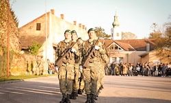 Soldaten in Militäruniformen marschieren in einer Parade vor einer großen Menschenmenge und Gebäuden. Der Himmel ist blau.