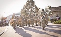 Eine Militärkapelle in Tarnuniformen marschiert mit Zuschauern am Bürgersteig die Straße hinunter. Bäume und ein Gebäude sind im Hintergrund zu sehen.
