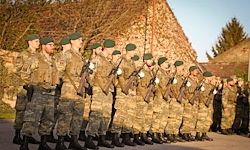 Eine Gruppe von Soldaten in Tarnuniformen steht in einer Reihe und hält Gewehre. Sie stehen vor einer Steinmauer, mit Bäumen im Hintergrund.