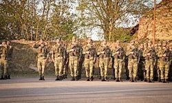 Eine Militäreinheit steht in Formation vor einer Steinmauer, unter klarem Himmel. Bäume sind im Hintergrund sichtbar.