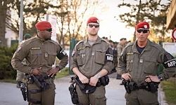 Drei Soldaten in Militäruniformen, jeder mit Sonnenbrille, Hut und Pistole, stehen auf einer Straße mit Gebäuden und Bäumen im Hintergrund.