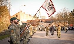 Soldaten in Uniform stehen in Formation mit einer Flagge vor einem Gebäude. Ein Mann in einem Anzug geht die Straße entlang.