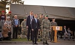 Eine militärische Zeremonie mit einem Mann in Anzug und zwei Militärmännern, einer in Grün und einer in Schwarz, vor einem Podium mit einer versammelten Menge.