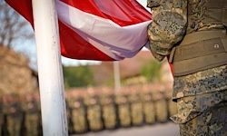 Ein Soldat in Militäruniform steht neben einem Fahnenmast bei einer militärischen Zeremonie, mit vielen anderen Soldaten, die hinter ihm in der Reihe stehen.