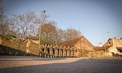 Eine Reihe von Militärangehörigen steht in Formation vor einem Steinwall und Gebäuden dahinter, unter einem klaren Himmel.