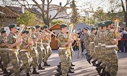 Eine Gruppe von Militärangehörigen in Tarnuniformen marschiert bei einer Parade mit Trompeten und Fahnen. Zuschauer beobachten sie an der Seite.