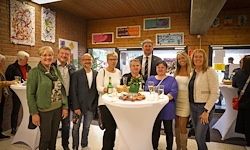 Eine Gruppe von Menschen steht um einen Tisch mit Essen, Getränken und einer grünen Pflanze herum und posiert für ein Foto. Sie befinden sich in einem Raum mit Kunstwerken an den Wänden.