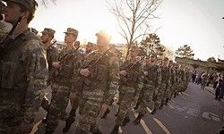 Eine Reihe von Soldaten in Tarnuniformen, die eine Straße hinuntermarschieren, mit Bäumen und Menschen auf dem Bürgersteig.