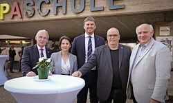 Fünf formell gekleidete Personen stehen gemeinsam vor einem weißen Tisch. Dahinter ist ein Gebäude mit dem Schild 'Passchull' zu sehen. Der Mann auf der rechten Seite trägt eine Brille und eine Uhr.