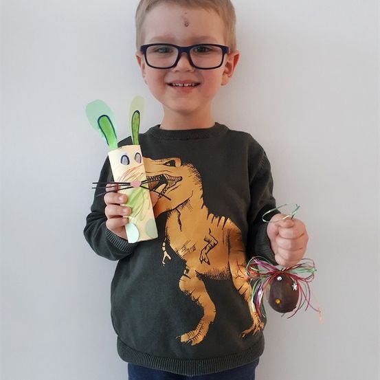 Ein junger Junge mit Brille und Dinosaurier-Shirt hält eine Papierbastelarbeit eines Kaninchens und ein dekoratives Osterei.
