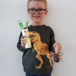 Ein junger Junge mit Brille und Dinosaurier-Shirt hält eine Papierbastelarbeit eines Kaninchens und ein dekoratives Osterei.