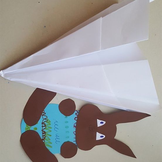 Ein brauner Papierhase mit blauen Augen und einem blauen Shirt ist an einem weißen Papierflugzeug auf einem hellbraunen Hintergrund befestigt.