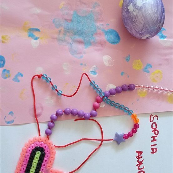 Eine bunte Perlenkette mit einem Herzanhänger und den Buchstaben L, A und N, auf einem rosa Hintergrund mit einem bemalten Ei und einer weißen Oberfläche.