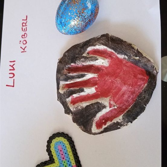 Ein blaues glitzerndes Ei, ein roter Handabdruck auf einem Stein und ein aus bunten Perlen gefertigter Buchstabe L liegen auf einem weißen Hintergrund. Die Namen 'Luki' und 'Kogerl' sind links in Rot geschrieben.