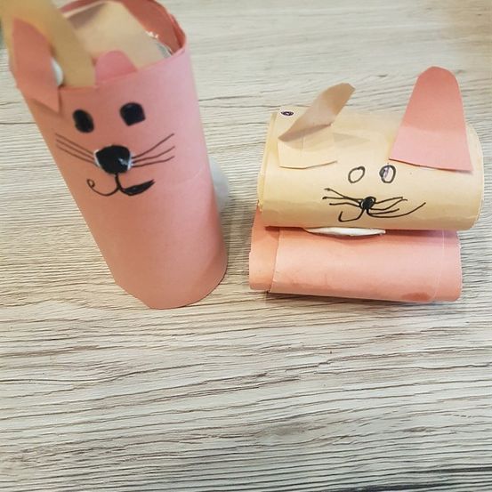 Zwei katzenförmige Bastelarbeiten aus Papierrollen liegen auf einer Holzoberfläche. Eine ist rosa mit einem gezeichneten Gesicht, die andere ist beige mit einem rosa Ohr.