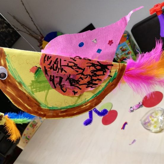 Ein farbenfrohes Papierbasteln eines Vogels mit einem rosa Schnabel und Schwanzfedern, die an einem Faden hängen, mit einer Nachricht darauf. Der Hintergrund zeigt einen Tisch mit verschiedenen bunten Objekten.