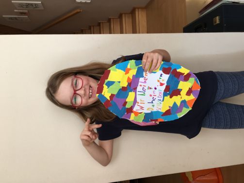 Ein junges Mädchen mit Brille hält eine farbenfrohe Papiercollage mit den Worten 'Wir lieben' und 'Daheim'. Sie lächelt und posiert für ein Foto.