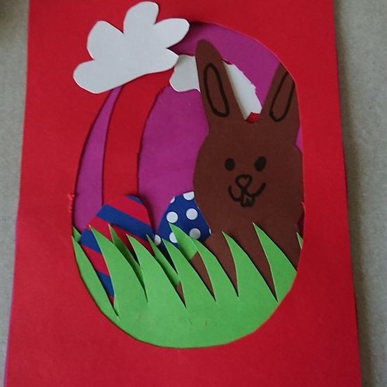 Eine handgefertigte Osterkarte mit einem braunen Hasen, der aus einem mit bunten Eiern gefüllten Osterkorb auf einem grünen Grashintergrund herausschaut.