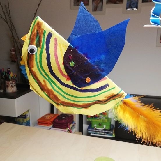 Ein bunter Papiervogel mit blauen Flügeln und einem gelben Körper, der an einer Schnur hängt. Der Vogel hat ein weißes Auge und eine gelbe Schwanzfeder. Der Hintergrund zeigt einen Raum mit einem Schreibtisch, einer Pflanze und verschiedenen Gegenständen.