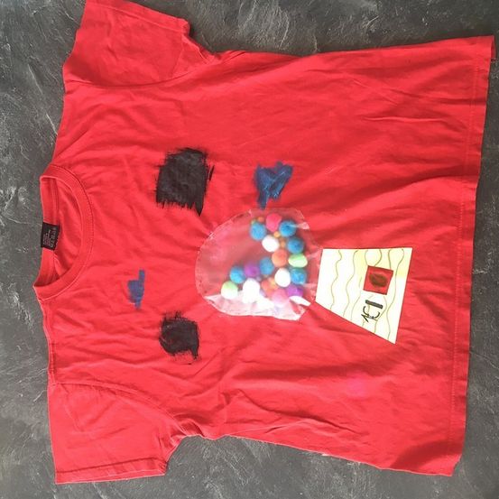 Ein rotes T-Shirt mit einem Kaugummiautomaten-Design auf der Vorderseite. Der Kaugummiautomat besteht aus durchsichtigem Kunststoff und ist mit bunten Kaugummis gefüllt. Auf dem Shirt befinden sich schwarze Farbspritzer.