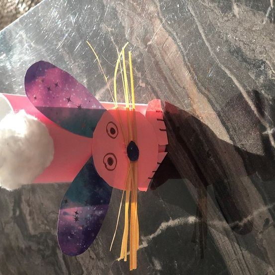 Eine rosa Papierbastelarbeit, die einem Hasen mit Flügeln und einem galaktischen Design auf den Ohren ähnelt. Es hat einen Wattebausch auf dem Kopf und steht auf einer Marmoroberfläche.