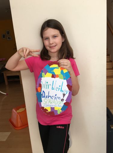 Ein junges Mädchen in einem rosa T-Shirt hält ein buntes Papierei mit den Worten 'Wir bleiben Daheim' und 'Schadwore Corona-Kreat'. Sie steht in einem Raum mit einer weißen Wand und einer hölzernen Treppe im Hintergrund.