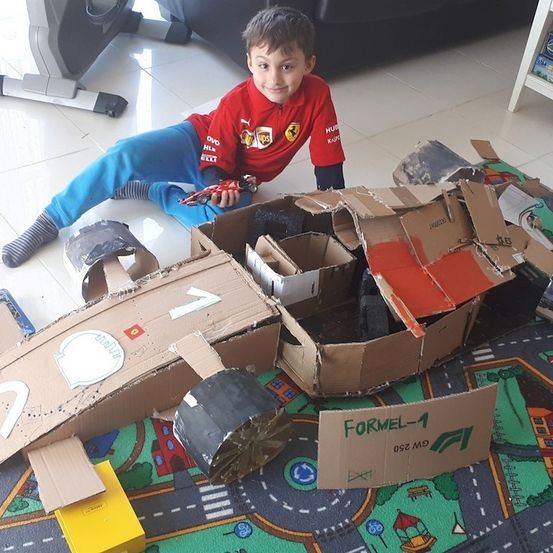 Ein junger Junge sitzt neben einem selbstgebauten Kartonauto auf dem Boden und hält ein Spielzeugauto in den Händen.