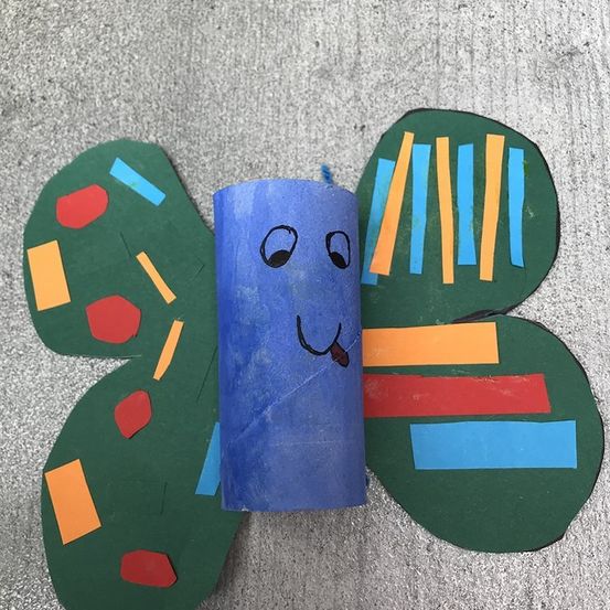 Ein handgefertigter Schmetterlingsbastel mit einem blauen Körper und bunten Flügeln, mit einem lächelnden Gesicht auf dem Körper. Die Flügel sind mit verschiedenen farbigen Papierstücken verziert.