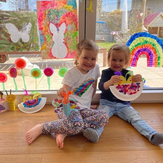 Zwei kleine Kinder sitzen vor einem Fenster auf dem Boden und halten Papierbasteleien in den Händen. Das Fenster ist mit bunten Papierausschnitten von Blumen, einem Regenbogen und einem Schmetterling verziert.