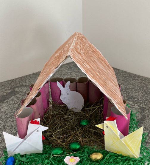 Eine handgefertigte Krippe aus Papierröhren und Bastelmaterialien, mit einem Kaninchen, zwei Hühnern und Ostereiern in einem Nest.