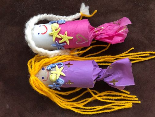 Zwei meeresjungfrauen-bastelarbeiten aus Papier und Garn, eine mit einem rosa-weißen Farbschema und die andere mit einem lila-weißen Farbschema, beide mit Seestern- und Muscheldekorationen.