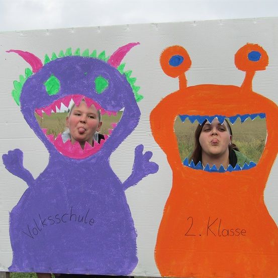 Zwei Kinder posieren in Ausschnitten von Monstern auf einem Papphintergrund. Das lila Monster hat die Worte 'Volksschule' darauf geschrieben, und das orange Monster hat '2. Klasse'. Beide Kinder stecken ihre Zungen heraus.