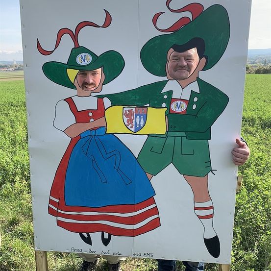 Zwei Personen stehen hinter einem bemalten Schild auf einem Feld. Das Schild zeigt einen Mann und eine Frau in traditioneller bayerischer Kleidung, wobei der Mann einen Schild hält.