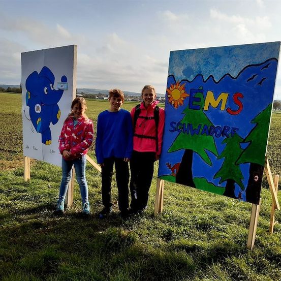 Drei Kinder stehen vor zwei großen bemalten Tafeln in einem Grasfeld, wobei eine Tafel einen blauen Elefanten und die andere eine farbenfrohe Landschaft mit dem Wort EMS zeigt.