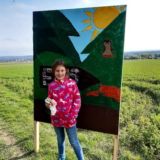 Ein kleines Mädchen steht auf einem Feld neben einem großen bemalten Brett mit einer Sonne und einer Eule darauf. Sie hält ein Sandwich und lächelt.