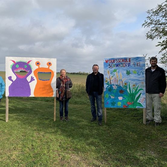 Vier Personen stehen auf einem Feld mit zwei bunten Postern hinter ihnen. Das linke Poster zeigt zwei Monstergesichter, während das rechte Poster eine Landschaft mit dem Text 'Volksschule Schwadorf 4.KL.' hat.