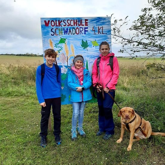 Drei Kinder und ein Hund posieren für ein Foto vor einem farbenfrohen Schild, das 'Volksschule Schwadorf 4.KL' besagt. Die Kinder tragen Jacken und der Hund ist an der Leine. Im Hintergrund ist ein grasiges Feld mit einem Baum und einem bewölkten Himmel zu sehen.