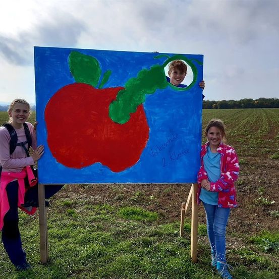 Zwei Mädchen stehen neben einer großen Malerei eines roten Apfels, aus dem ein Junge herausschaut. Das Gemälde steht auf einem Holzständer in einem Feld.