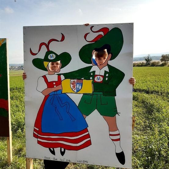 Zwei Kinder stehen hinter einer großen Pappaufsteller-Karikatur eines Mannes und einer Frau in traditioneller bayerischer Kleidung, die einen Schild mit einem Wappen hält. Sie befinden sich in einem Grasfeld mit Bergen in der Ferne.