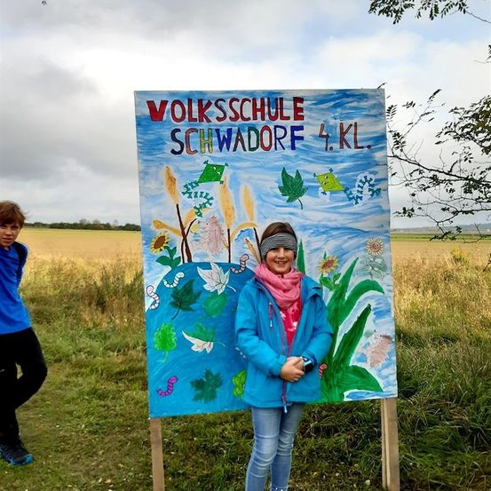 Ein Mädchen in einer blauen Jacke steht vor einem farbenfrohen Schild mit den Worten 'Volksschule Schwadorf 4.KL.' auf einem Grasfeld.