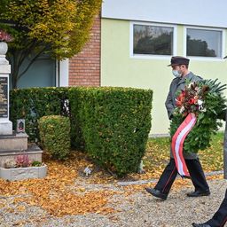 Zwei Männer in Uniform und Masken gehen an einem Denkmal vorbei und halten einen Kranz mit roten und weißen Bändern.