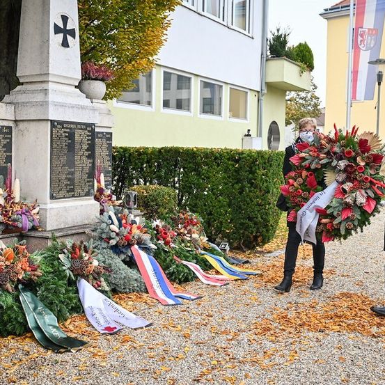 Zwei Personen in formeller Kleidung und Masken stehen neben einem Denkmal, das mit Kränzen und Flaggen geschmückt ist. Das Denkmal hat ein Kreuz und eine Tafel mit Namen und Daten.
