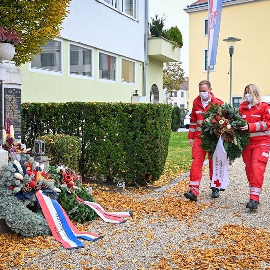 Zwei Personen in roten Uniformen und Gesichtsmasken gehen an einem Denkmal vorbei, das mit Kränzen und Flaggen geschmückt ist. Hinter ihnen ist ein Gebäude mit einer Flagge und einer Hecke zu sehen.