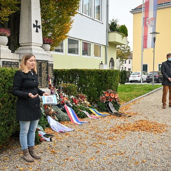 Eine Frau in einem schwarzen Mantel steht vor einem Denkmal mit Blumen, einer Flagge und einem Kreuz auf einem Steinpfeiler. Ein Mann mit Maske steht hinter ihr auf einem Kiesweg.