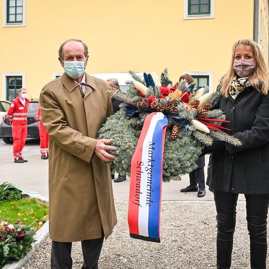 Ein Mann und eine Frau mit Gesichtsmasken stehen vor einem Kranz mit einem Band, auf dem 'Marktgemeinde' steht. Dahinter stehen zwei Personen in roten Uniformen neben einem Auto.